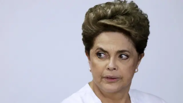 Dilma com olhar sério olhando para o lado, vestindo branco ehttps apostasesportiva vipfrente a painel branco