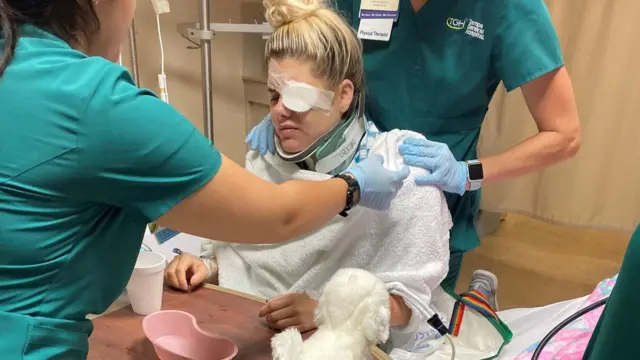 Oto u bolnici sa zavojima i povezom na oku, okružena medicinskim osobljem koje joj pomaže u svakom pokretu