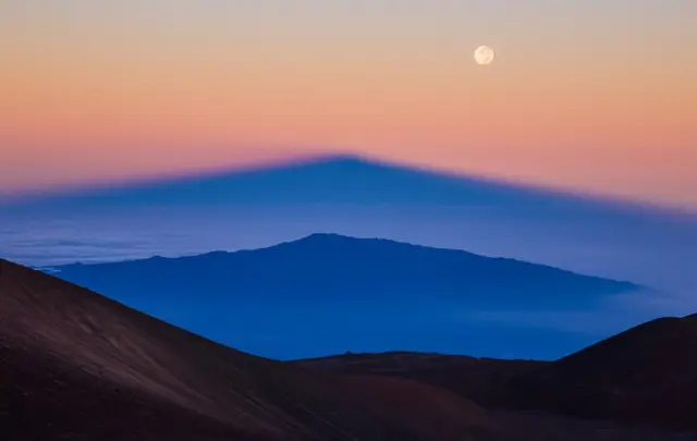 Тень Мауна-Кеа (Гавайи), проецируется восходящим солнцем над спящим вулканом Хуалалаи, в то время как на небе сияет луна