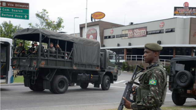 Homem fardado com armamento pesado em rua, com veículo repleto de outros soldados atrás