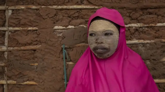 A girl wearing a clay mask and pink scarf smiles.