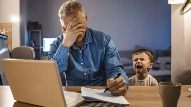 Un homme la tête dans les mains devant un ordinateur portable alors qu'un enfant pleure