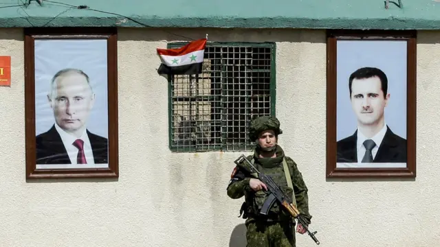 جندي من الجيش الروسي يقف خارج بناية في سوريا عليها صورتين لفلاديمير بوتين وبشار الأسد