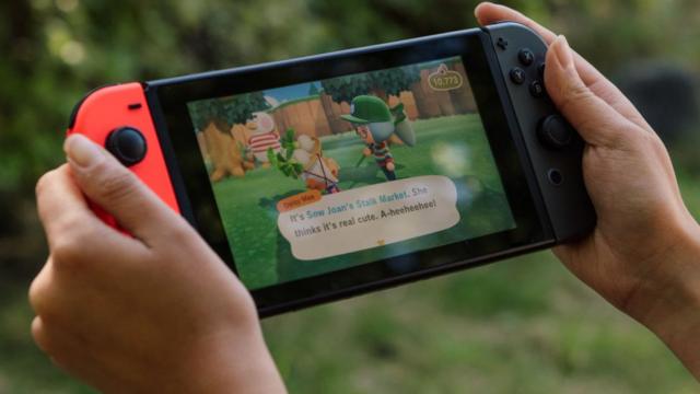 A pair of hands holds a Nintendo Switch console in an outdoor - garden - environment. On the screen is a shot from Animal Crossing - two characters are in conversation, as denoted by an on-screen dialogue box.