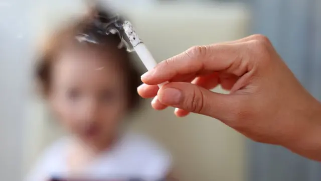 Pessoa segura cigarro com a mão com uma criança ao fundo