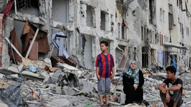 जुलाई 2014 में ग़ज़ा पर हुई बमबारी में ध्वस्त हुई इमारत के सामने एक परिवार