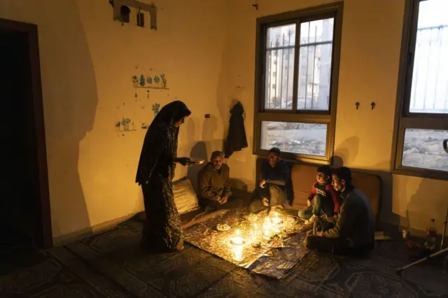 3 رجال وطفل وامرأة على مائدة إفطار بسيطة دون كهرباء، الغرفة مضاءة بعدد من الشموع.