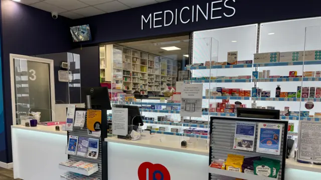 Un mostrador de farmacia con un gran cartel que dice "MEDICAMENTOS". Filas de medicamentos envasados ​​se encuentran en los estantes detrás del mostrador.