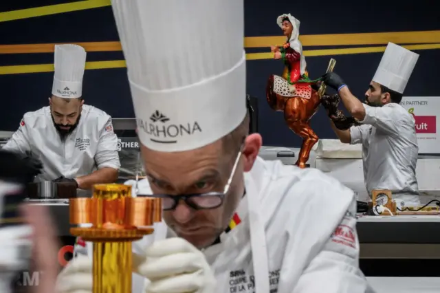Three men in chef's hats and gowns work on intricate pasty dishes.
