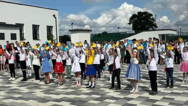 На передньому плані - молодші школярі у святковому одязі з піднятими догори руками - одна рука в кожного жовта, інша -синя