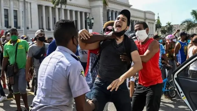 Homens (não fardados) seguram manifestante, que gritano funciona bwinfrente a policial na rua