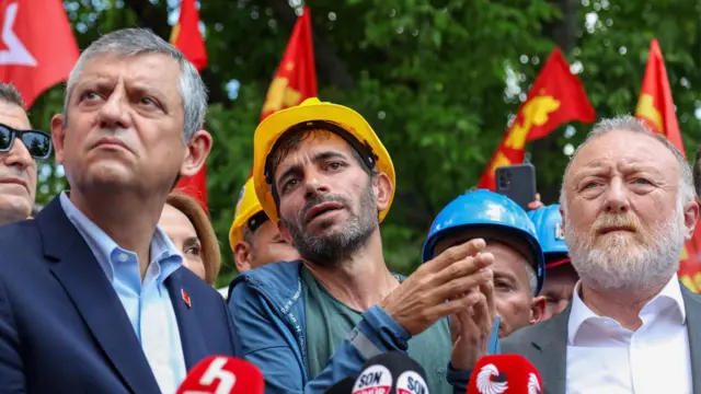 Saroı kasklı bir madenci konuşuyor, önünde mikrofonlar var. Yanında Özgür Özel duruyur