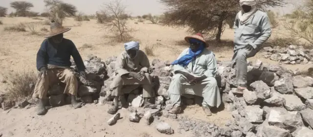 Le maitre maçon Maouloune Baigna Badou, en turban bleu, portant un tingade, assis avec son équipe sur un site d'extraction du calcaire Alhor.