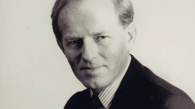 Una fotografía en blanco y negro de John Smyth con una camisa a rayas y un traje oscuro.