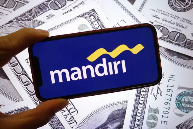 Dalam ilustrasi foto ini, logo perusahaan Bank Mandiri terlihat ditampilkan di layar ponsel cerdas dengan uang kertas dolar Amerika Serikat di latar belakang.