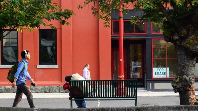 Imagem mostra dois homens caminhando na rua, enquanto outro, sem teto, está inclinado sobre um banco de praça em Portland (EUA)