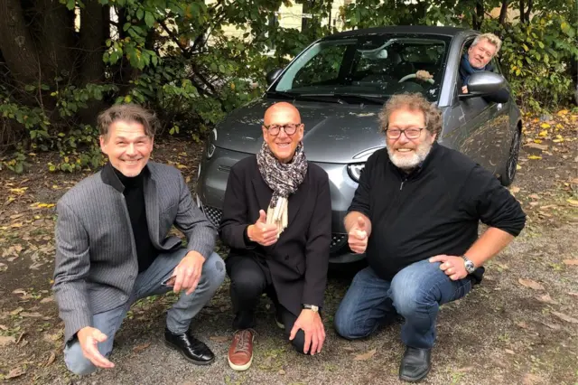 Três homens brancos idosos posambet pt slotsfrente a um carro cinza, outro homem está dentro do carro