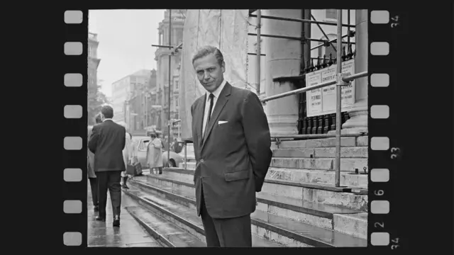 Fotografía en blanco y negro de David Attenborough con traje frente a unas escalinatas urbanas, con andamios y peatones al fondo, enmarcada como negativo de película.