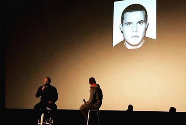 John McAvoy speaking at a public event, with his 2005 mugshot on screen behind him