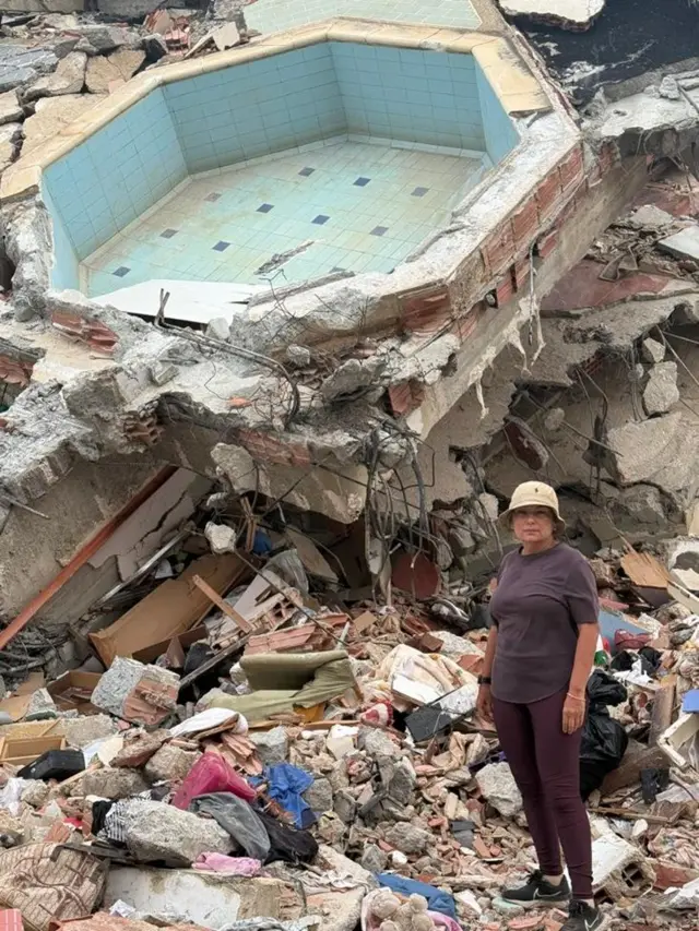 María Lourdes Pérez, vestida con un pantalón y camisa marrones y un gorro beige, está parada en una zona llena de escombros. Detrás de ella está lo que parece ser una piscina pequeña, piedras, hormigón, partes de una edificación, muebles destruidos, ropa.
