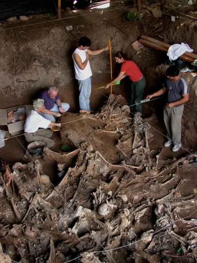 Hombres y mujeres excavando en un lugar lleno de esqueletos que están medio sepultados.