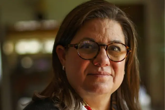 Laurie Ann Ximénez-Fyvie, uma mulher branca, de cabelos castanhos e que usa óculos marrons, olha para a câmera sorrindo