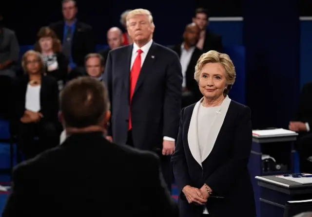 Capres Amerika Serikat dari kubu Partai Demokrat saat itu, Hillary Clinton, tengah mendengar pertanyaan salah seorang penonton debat yang merupakan perwakilan masyarakat, sementara Donald Trump yang merupakan lawannya turut mendengarkan di belakangnya.