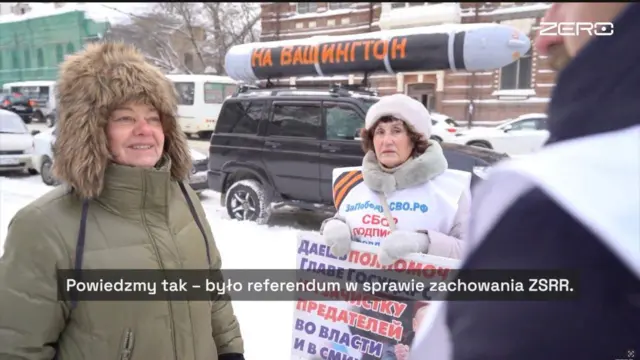 На заснеженной улице стоят несколько человек, пожилая женщина в светлом пуховике с мехом держит плакат с надписью на русском языке: «Даешь полномочия главе государства на зачистку предателей во власти и СМИ». Сзади виден автомобиль с муляжом ракеты. На переднем плане – Верниковская, которая разговаривает с участником акции. В нижней части кадра субтитр: «Скажем так – был референдум о сохранении СССР».