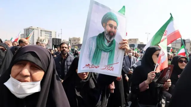 黒衣の女性たちが、イラン国旗や最高指導者の肖像画などを手に道を歩いている。後ろには男性たちも歩いている