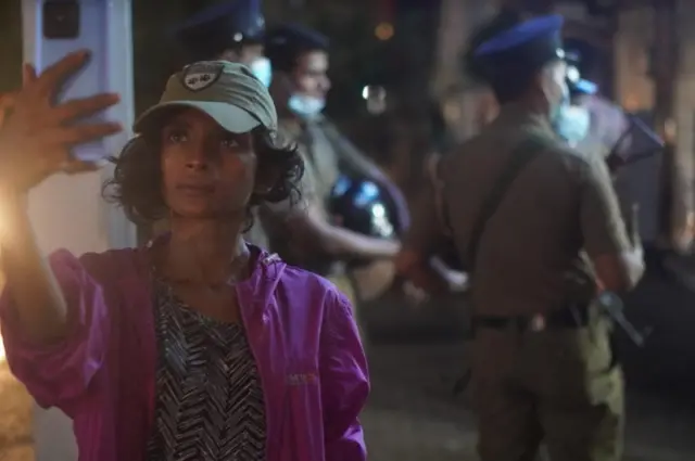 Melani Gunathilaka filming from her phone with police behind her