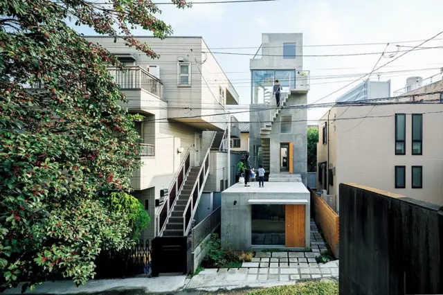 Vivienda urbana de hormigón visto con terrazas escalonadas y escalera exterior, rodeada de edificios residenciales.