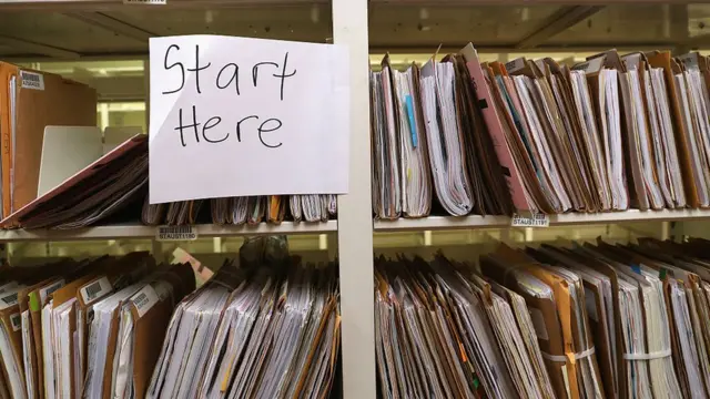 Una oficina del USCIS con estantes llenos de expedientes y un papel que dice "Start here" ("Empiece aquí")