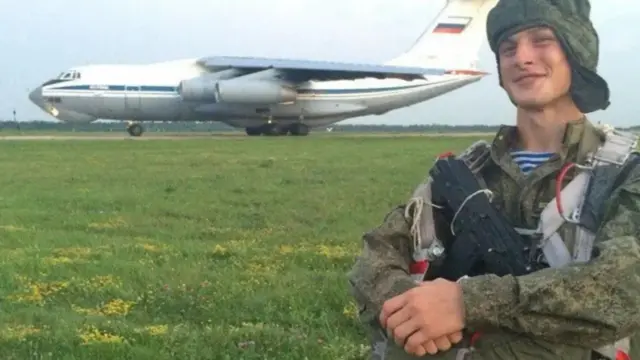 Жанговар кийимда қўлларини бирлаштириб турган ёш йигит ҳарбий самолёт олдида туриб, камерага табассум қиляпти.