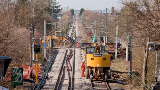 Se están construyendo nuevas vías del metro sobre un camino de grava que se pierde en el horizonte. La vía está rodeada de árboles y delimitada por estructuras metálicas grises que sostienen el cableado aéreo. Hay dos vehículos de trabajo amarillo brillante en la vía y un par de trabajadores con uniformes naranjas de alta visibilidad.