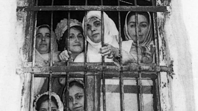 Şenler'in Bursa Cezaevi'ndeki bir fotoğrafı. Demir parmaklı bir pencereden bir grup başka kadınla birlikte bakıyor.