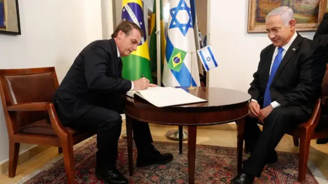 Bolsonaro e Netanyahu sentados à mesa; o presidente brasileiro assina documentos