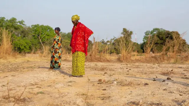 Deux femmes se tiennent debout sur un terrain sec, aride et poussiéreux, inspectant les environs, avec de hautes herbes desséchées derrière elles et des arbres verts au loin.