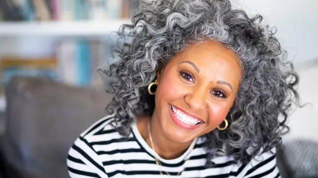 一位女性坐在室內的沙發上，對著鏡頭微笑。她有捲曲的及肩灰白髮，穿著黑白條紋T恤，戴著兩條金項鍊和金色圈形耳環。她身後是一個書櫃。。