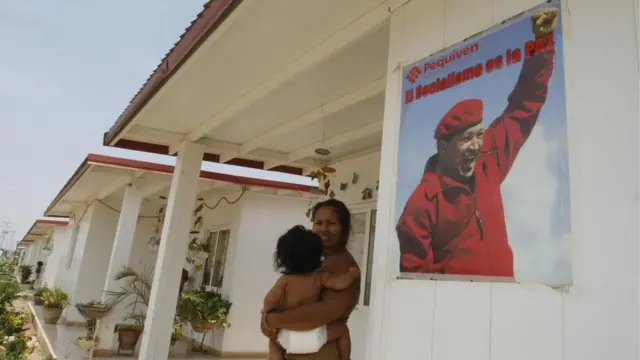 Una mujer frente a una casa con un cartel de Hugo Chávez al lado