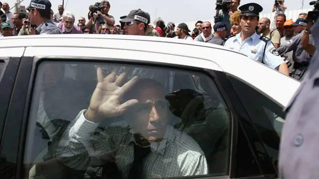 Israeli whistleblower Mordechai Vanunu leave prison for a white car, wearing a chequered shirt and black tie, wit im hand on di car window, as crowds of journalists and police officers surround im car