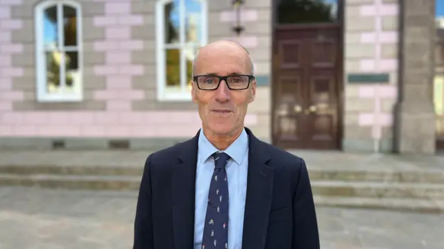 El diputado Jonathan Renouf, un hombre calvo con gafas oscuras rectangulares, lleva una camisa azul claro, una corbata azul marino y un traje azul oscuro. Mira fijamente a la cámara, de pie frente a la Cámara de Representantes de Jersey. Se ve una puerta doble de madera por encima de su hombro, a la derecha de la imagen.