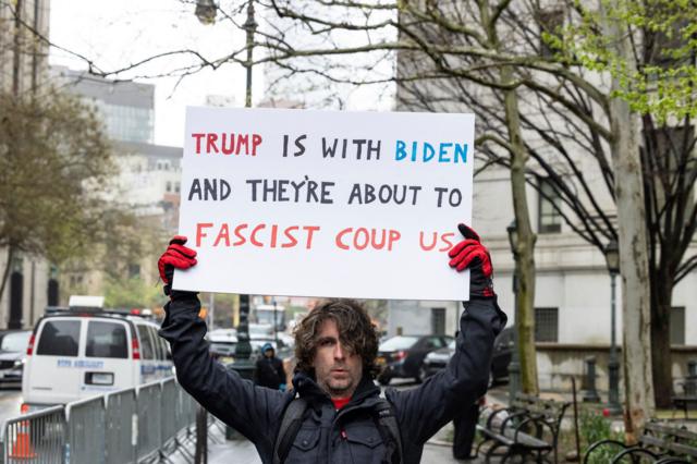 Максвелл Азарелло держит плакат с надписью «Трамп и Байден заодно, они готовят фашистский переворот»