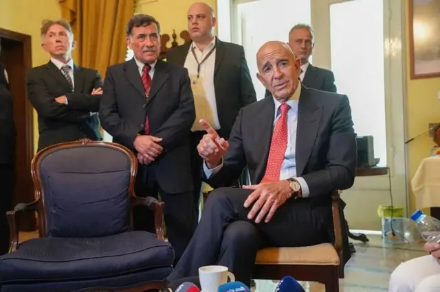 tom barrack bir sandalyede oturmuş konuşurken arkasında da bazı görevliler ayakta duruyor