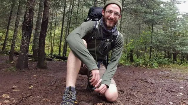Alex dey pictured in woodland, e wear shorts and walking boots while kneeling on di ground