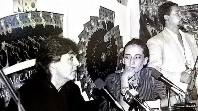 Em foto preto e branco, Paul McCartney fala no microfone, observado por May Waddington