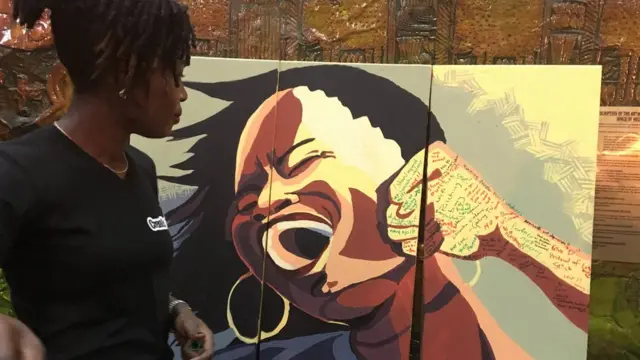 A girl dey stand beside painting of woman wey dem dey beat.