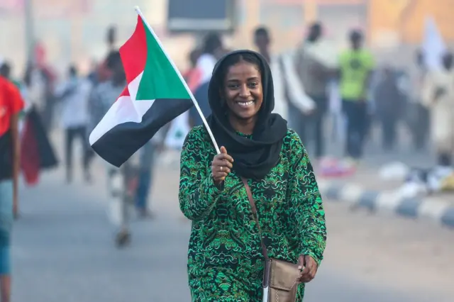 A smiling woman standing in the street holds a Sudanese flag.