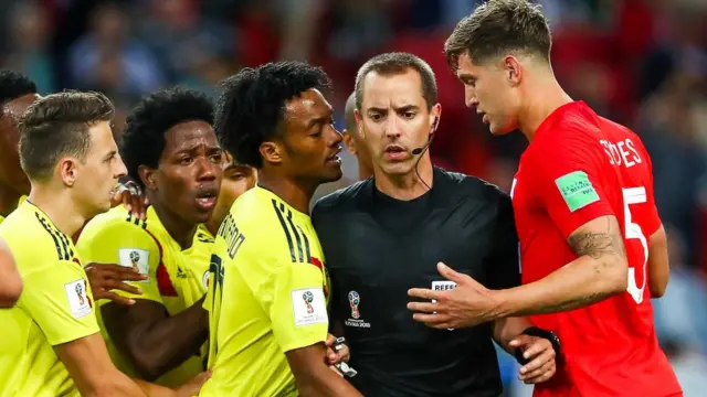 John Stones and referee Mark Geiger are surrounded by Colombia players