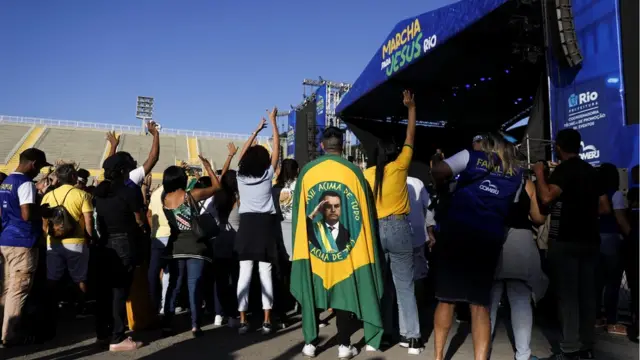 Fotografia colorida mostra um homemcomprar roleta de cassinocostas enroladocomprar roleta de cassinouma bandeira do Brasilcomprar roleta de cassinoque no centro há uma fotocomprar roleta de cassinoBolsonaro; o homem está ao ladocomprar roleta de cassinoalgumas pessoascomprar roleta de cassinofrente a um palco onde está escrito "Marcha para Jesus Rio"