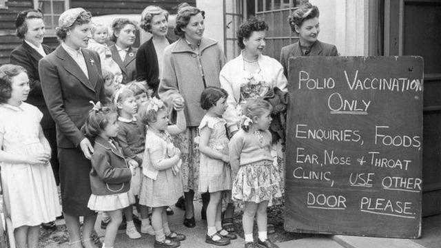 Crianças e mulheres fazem fila do lado de fora de um centro de vacinação. com uma placa dizendo: "Apenas vacinação contra poliomielite"